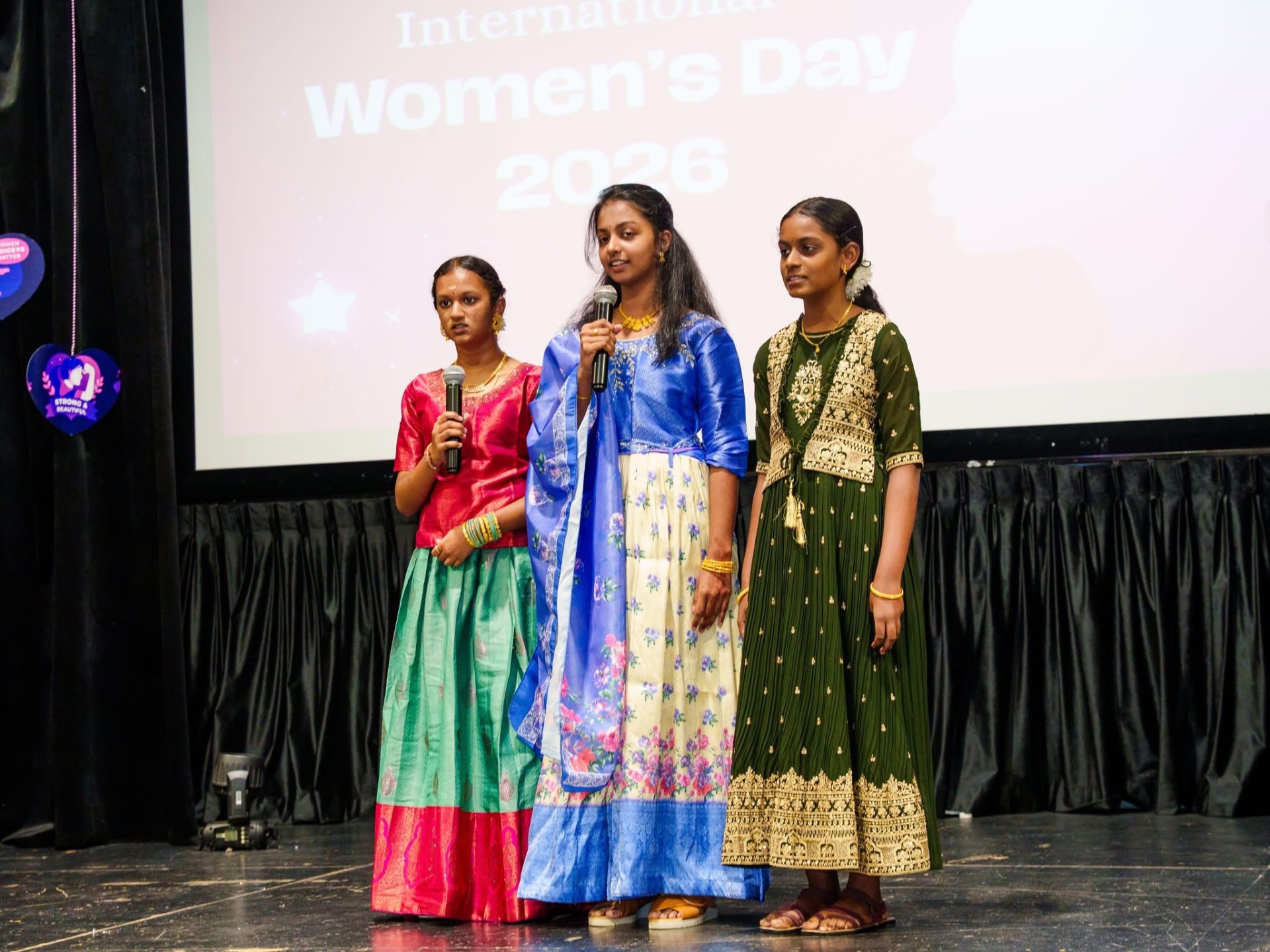 Three young hosts speaking on stage at International Women's Day 2026